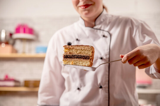 Hand holding a slice of layered chocolate sponge cake on a server, with decorated desserts in a pastry workshop.