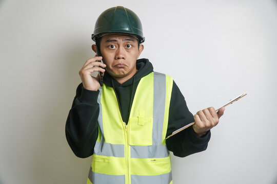 Concerned Asian Construction Worker Talking on Smartphone, Holding Clipboard