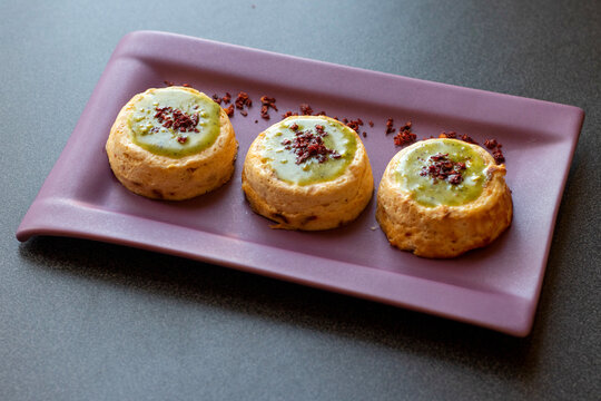 Three baked ricotta timbales topped with creamy pistachio pesto and crumbled crispy red pepper, arranged on a rectangular purple plate, overhead angled view.