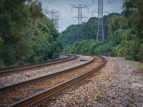 Curving railway tracks winding through a green forest with tall metal electrical transmission towers under an overcast sky