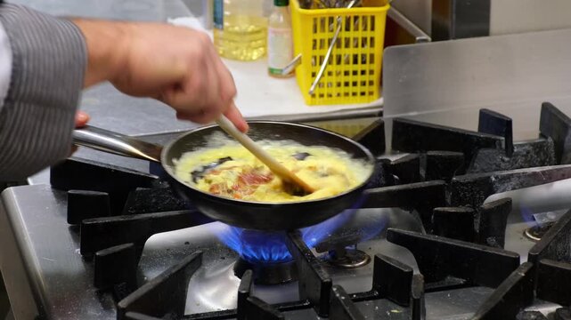 Chef hands stirring eggs and ingredients in hot pan on open gas stove burner flame in restaurant kitchen. Cooking scrambled omelette breakfast close-up.