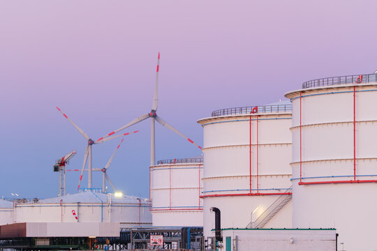 Large oil storage tanks at an industrial refinery facility with wind turbines in the background. Petroleum fuel storage tanks near a green energy wind farm.