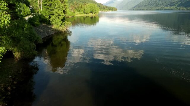 Calm lake mirrors sky and trees perfectly. Still water reflects clouds and mountains. Green forest borders shoreline with rocky edges. Peaceful nature scene perfect for relaxation or travel projects