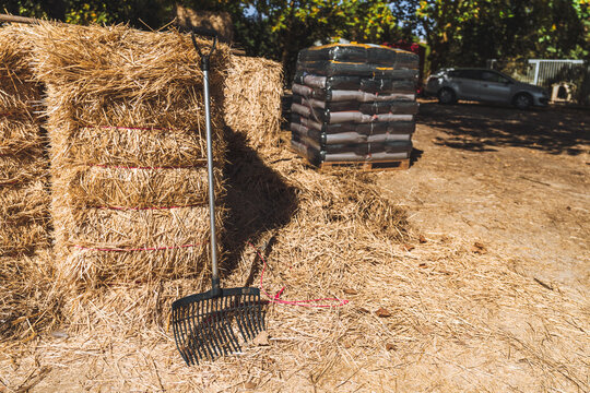 Black bedding fork rests against stacked golden hay bales at an outdoor stable work area, with wrapped feed bags and a parked car blurred behind