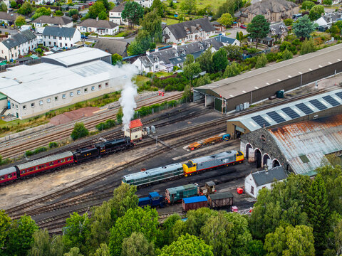 Aerial view of the historic Aviemore railway station with a steam locomotive emitting smoke, diesel trains in the depot, and town buildings in Aviemore, Scotland, United Kingdom.