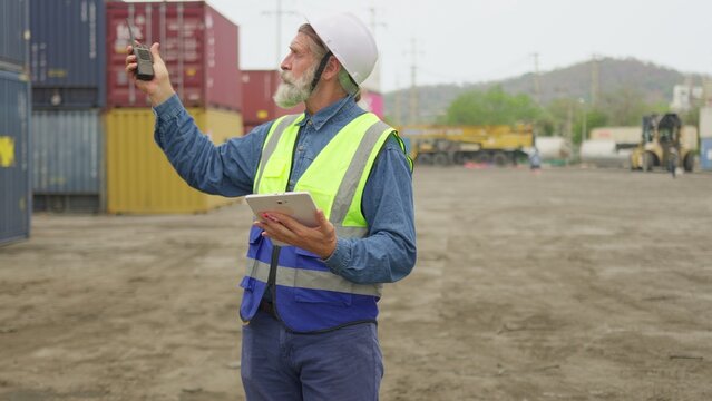 Senior male foreman communicating on a walkie-talkie and holding a tablet to manage freight logistics and oversee operations in a cargo container yard.