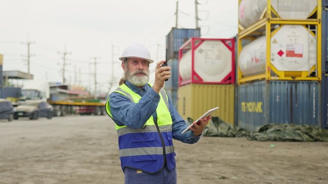 An experienced logistics supervisor coordinates freight operations using a digital tablet and walkie-talkie in a large commercial port with stacked intermodal containers.