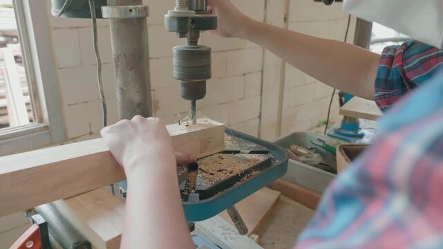 A close-up of a person using a drill press to create a hole in a wooden block. The workshop setting highlights the woodworking process and tools in action, showcasing craftsmanship and precision in cr