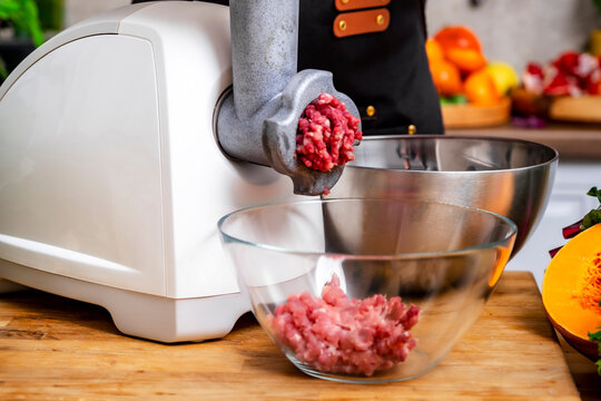 Woman uses electric meat grinder, mincing raw pork into glass bowl. Domestic kitchen food equipment culinary concept.