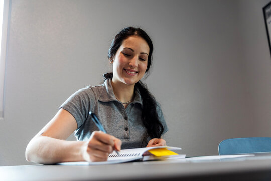 Smiling woman writing in a notebook at a desk in a bright room.
