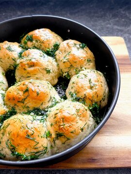 Freshly baked round bread , pampushki, garlic rolls topped with herbs arranged in a black bowl on a wooden cutting board against a dark stone surface