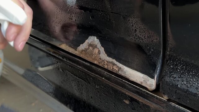 Close-up of a hand using sandpaper to remove rust from a car door trim, showing the gradual cleaning process with visible dirt and moisture on the surface