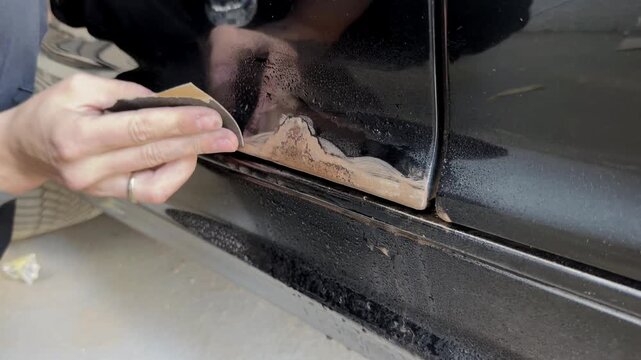 Close-up of a hand using sandpaper to remove rust from a car door trim, showing the gradual cleaning process with visible dirt and moisture on the surface