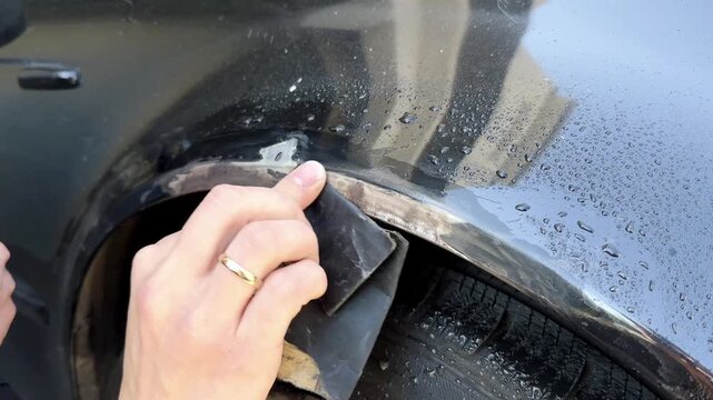 Close-up of a hand using sandpaper to remove rust from a car door trim, showing the gradual cleaning process with visible dirt and moisture on the surface
