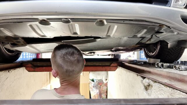Male auto mechanic inspecting car components from beneath vehicle on a lift, showcasing detailed actions in a garage environment with tools and equipment visible