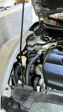 Male mechanic in beige shirt inspects car engine components under the hood in a garage, with tools and spare parts visible in the background