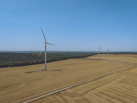 Aerial view of wind turbines standing in a vast golden wheat field with a forest border under a clear blue sky in Saint-Amand-sur-Fion, Grand Est, France.