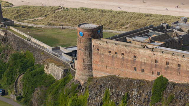 Aerial view of Bamburgh Castle with its historic stone walls, clock tower, and surrounding grassy dunes overlooking the coast in Bamburgh, England, United Kingdom.