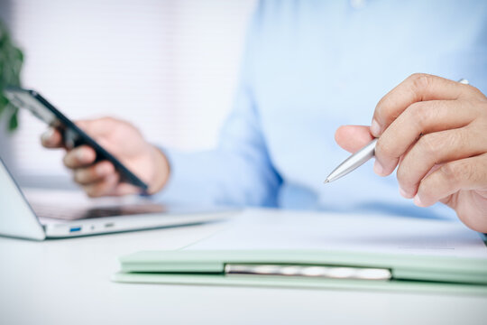 Businessman holding pen ready to sign contract document and using smartphone at office desk, concept of business agreement, financial planning, corporate work, and communication.