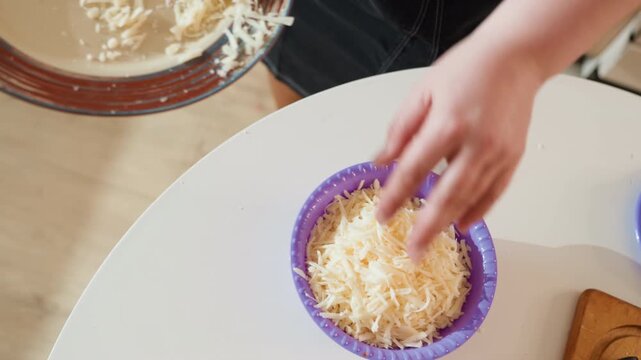 Overhead kitchen view showing hands pouring shredded cheese from mixing bowl into purple plastic colander on round white tabletop, capturing simple cooking preparation and gentle movement