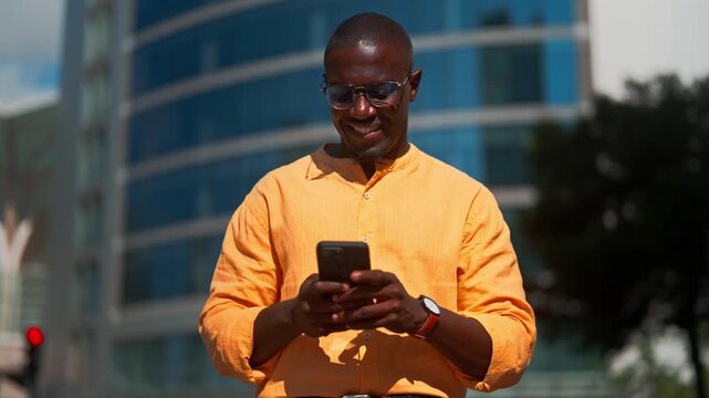 Black businessman using smartphone, texting and smiling, standing outdoor near office building. African american business man works as entrepreneur, checking messages outdoors, managing accounts.
