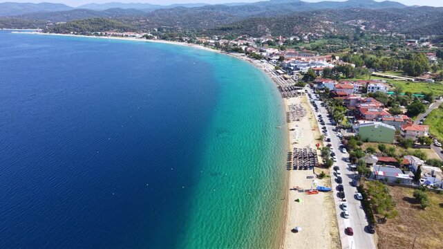 Aerial view of Toroni in Sithonia, Halkidiki, Greece, showing a busy Mediterranean beach with turquoise crystal-clear water, swimmers, sunbeds and beachfront hotels on a sunny summer day.
