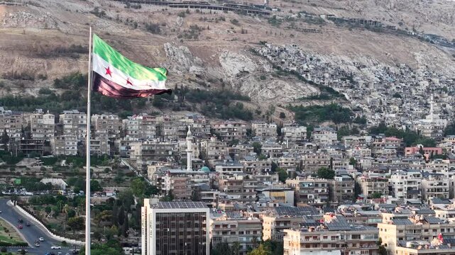 A scenic drone view shows Umayyad Square in Damascus bathed in sunset light as a massive Syrian flag waves over the landmark plaza and surrounding city landscape.