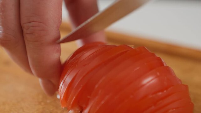 Tight kitchen scene shows hand slicing ripe tomato into thin even pieces on wooden board, with careful knife control, fresh red texture, warm light, and precise food preparation, with tomato, knife