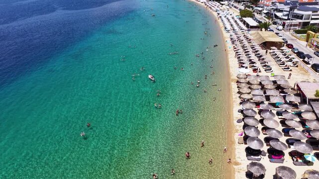 Aerial view of Toroni in Sithonia, Halkidiki, Greece, showing a busy Mediterranean beach with turquoise crystal-clear water, swimmers, sunbeds and beachfront hotels on a sunny summer day.