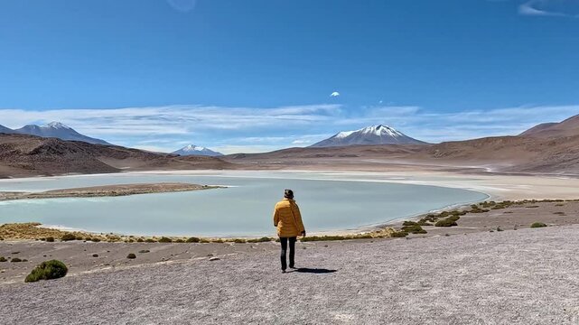 Young woman in a yellow jacket walking downhill towards a beautiful high altitude lagoon, surrounded by mountains and volcanoes in bolivia