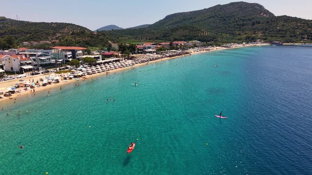 Aerial view of Toroni in Sithonia, Halkidiki, Greece, showing a busy Mediterranean beach with turquoise crystal-clear water, swimmers, sunbeds and beachfront hotels on a sunny summer day.