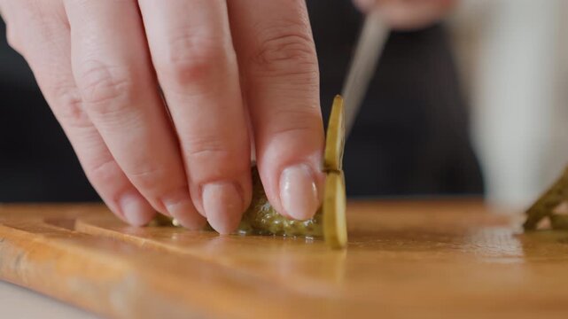 Tight low-angle close-up shows adult hands slicing small pickled green pepper on wooden cutting board, with sharp blade motion, glossy texture, warm light, and focused culinary preparation