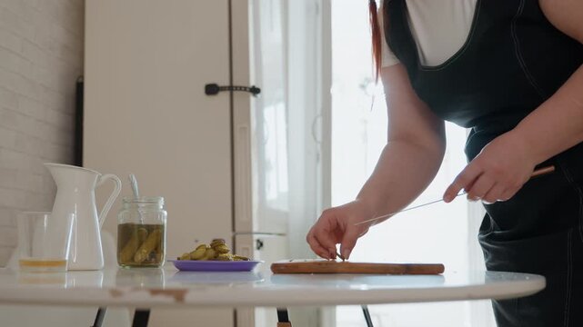 Middle aged woman bends forward beside round table, using fingertips to place or pick up small item while other hand steadies nearby, creating focused movement in sunlit kitchen interior