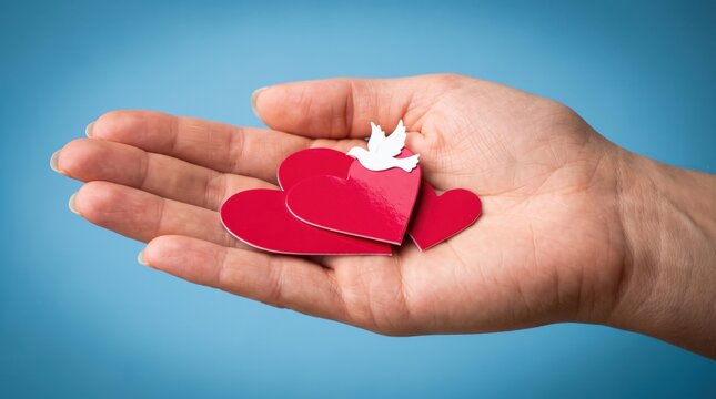 Human hand holding vibrant red paper hearts with small white dove cutout against a soft focused smooth blue studio background