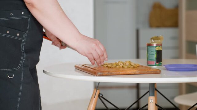 Close view of hands in dark apron finishing toppings on flatbread or thin crust pizza at round table in home kitchen, with pinching, sprinkling, pressing, and smoothing motions in soft daylight