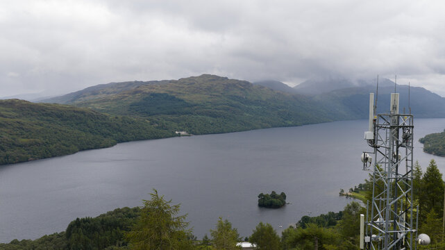 Aerial view of Loch Sloy with dark waters, a small island, and surrounding green hills under a cloudy sky, featuring a telecommunications tower in Argyll and Bute, Scotland, United Kingdom.