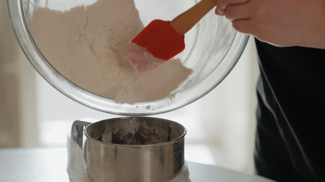 Close-up shows hands tipping glass bowl to pour flour into round metal sifter while orange spatula scrapes remaining flour, creating visible cascade and fine dust during baking preparation