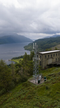 Aerial view of a communication tower situated on a grassy hillside overlooking the dark waters of Loch Sloy under a cloudy sky in Argyll and Bute Council, Scotland, United Kingdom.