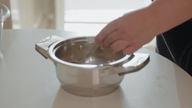 Close-up side view of hand moving metal colander inside matching bowl on bright kitchen countertop, with gentle water movement, droplets, shiny reflections, and soft natural daylight creating clean