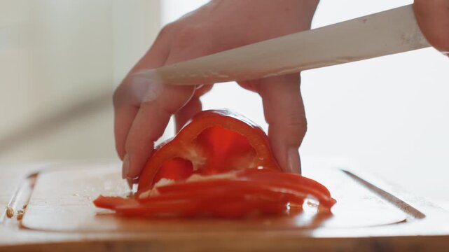 Close view of hands slicing ripe tomato into thin even rounds on wooden board, then stacking slices with fingertips beside resting knife in bright kitchen, showing careful food prep and fresh detail