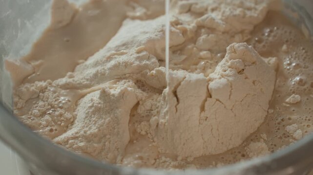 Overhead close-up of flour in stainless steel mixing bowl as thin stream of liquid pours into dry mound, creating darker damp patches, small bubbles, surface cracks, and clumps during dough