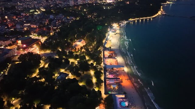 Top-down aerial night view of an illuminated seaside beach with restaurants, rows of sun loungers and umbrellas beside dark ocean waves, showing vibrant coastal nightlife and tourism.