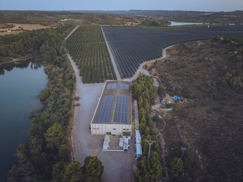 Aerial view of Finca El Pino featuring a large building with solar panels on the roof situated next to a river and expansive agricultural fields in Caspe, Aragon, Spain.