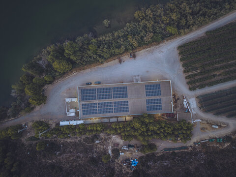 Aerial view of Finca El Pino featuring a building with solar panel arrays on the roof, surrounded by trees, agricultural fields, and a dirt road in Caspe, Aragon, Spain.