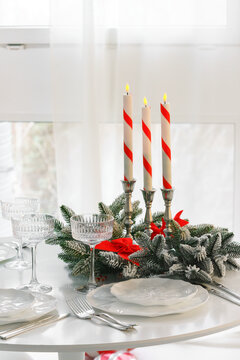Elegant Christmas table setting with red and white striped candles, evergreen branches, red bows, crystal glasses and white plates in a bright dining room on window background.