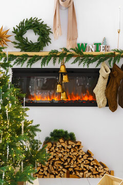 Cozy Christmas interior with a decorated fireplace, evergreen garland, wreath, stockings, bells, Christmas tree and stacked firewood. Festive living room, holiday mood, aesthetic decor