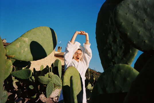 Woman Yawning Among Cacti With Arms Raised Under Blue Sky