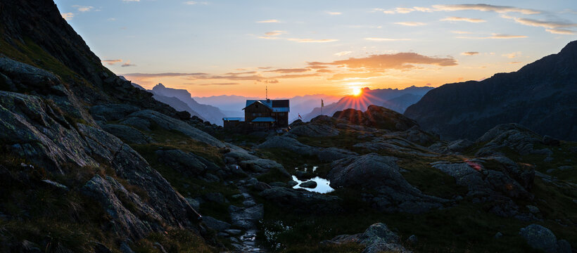 Bergh&uuml;tte in den Tiroler Alpen bei Sonnenaufgang mit aufgehender Sonne &uuml;ber alpiner Berglandschaft und kleinem Bergsee in der Morgend&auml;mmerung