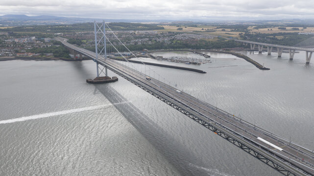 Aerial view of the Forth Road Bridge spanning the Firth of Forth with the Queensferry Crossing in the background, featuring steel suspension cables in South Queensferry, United Kingdom.