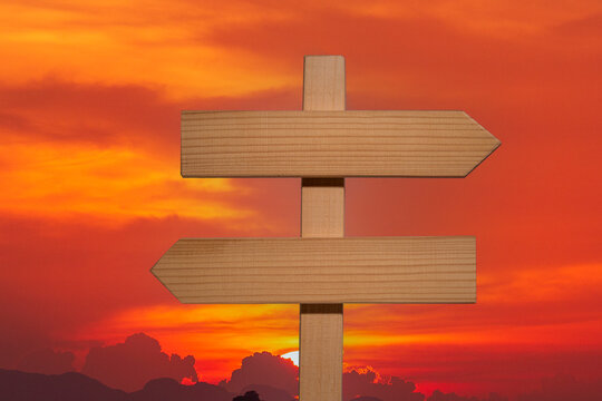 Blank wooden directional signpost against dramatic sunset sky. Empty wooden sign with two arrows pointing in opposite directions standing against bright orange and red sunset sky with clouds and sun